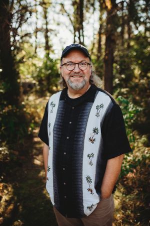 A man with glasses and a cap standing in the woods.