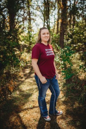 A woman standing in the woods wearing jeans and a maroon shirt.